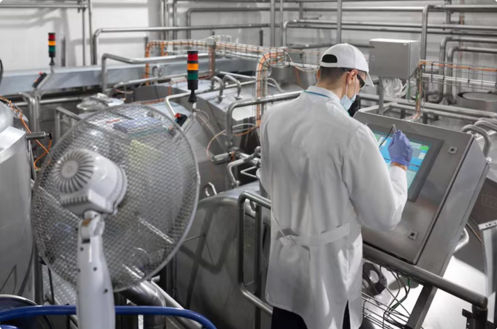 Operator in a white coat and cap using a touchscreen control panel on a stainless steel production line.