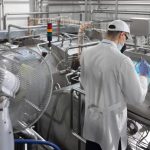 Operator in a white coat and cap using a touchscreen control panel on a stainless steel production line.