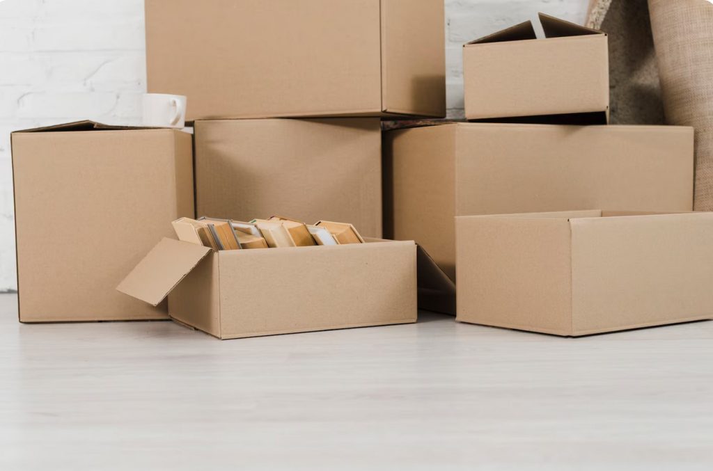 Stack of cardboard moving boxes stacked against a white wall, some sealed, with a mug resting on one box and packing materials visible nearby.