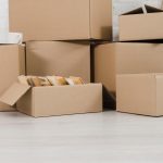 Stack of cardboard moving boxes stacked against a white wall, some sealed, with a mug resting on one box and packing materials visible nearby.
