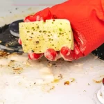 Gloved hand holding a yellow sponge and scrubbing a greasy stove top with soap suds nearby, showing a cleaning scene.