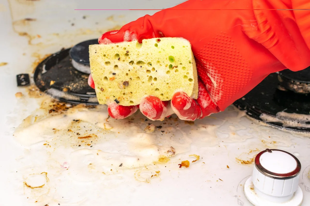 Gloved hand holding a yellow sponge and scrubbing a greasy stove top with soap suds nearby, showing a cleaning scene.