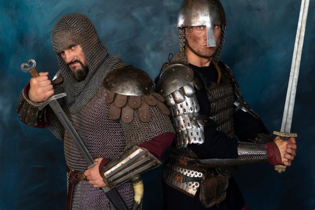 Two knights in full plate and chainmail armor pose with swords, standing side by side against a dark blue backdrop.