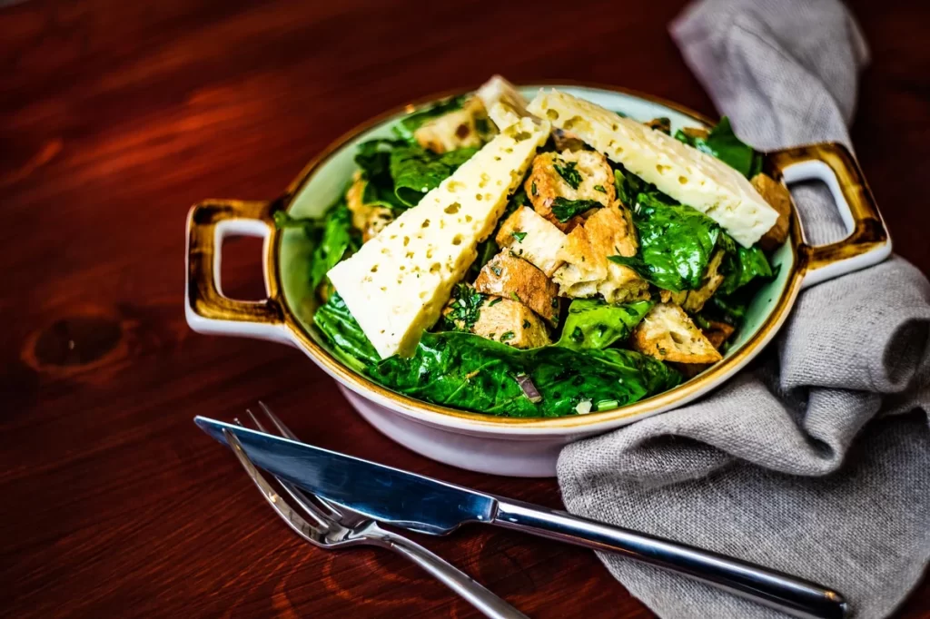 Mixed green salad in a shallow ceramic bowl, topped with Parmesan wedges and crunchy croutons on a dark wooden table.