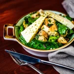 Mixed green salad in a shallow ceramic bowl, topped with Parmesan wedges and crunchy croutons on a dark wooden table.