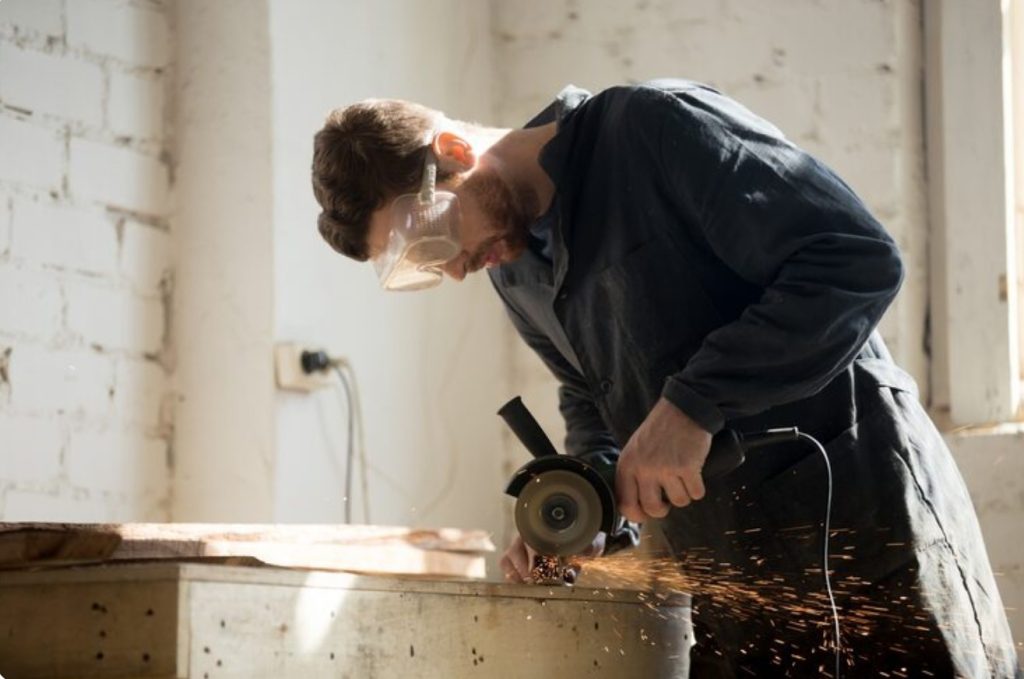 Man wearing safety goggles leans over a workbench, using a handheld grinder that sparks wood shavings onto the surface