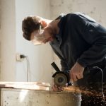 Man wearing safety goggles leans over a workbench, using a handheld grinder that sparks wood shavings onto the surface