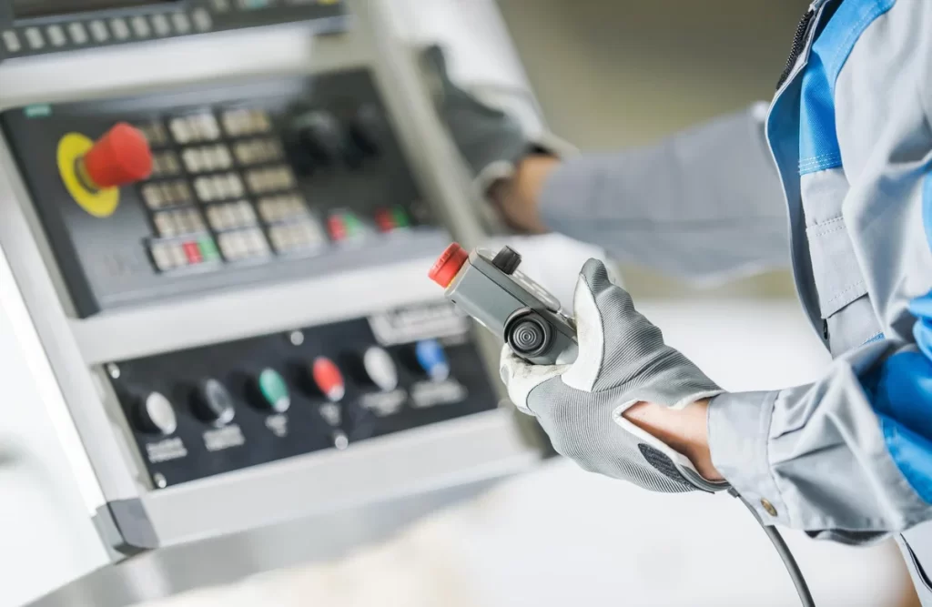 Worker in protective gloves uses a handheld control on an industrial machine panel with a red emergency stop button.