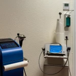 Clinical room with two medical devices: a blue touchscreen machine on the left and a smaller monitor with blue display on the right, plus a paper towel roll nearby.