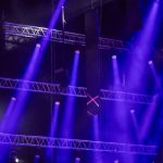 Concert stage with purple and blue spotlights and metal truss rigging overhead. A dark venue with lighting beams crisscrossing.