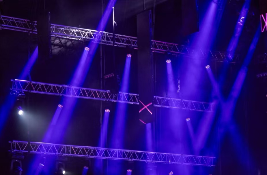 Concert stage with purple and blue spotlights and metal truss rigging overhead. A dark venue with lighting beams crisscrossing.